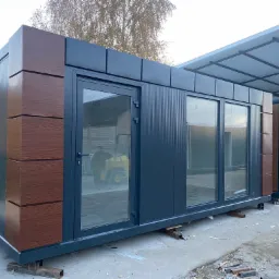 Gotowy modułowy pawilon handlowy z płyt warstwowych w kolorze grafitowym z drewnopodobnymi elementami dekoracyjnymi, osadzony na metalowych podporach.