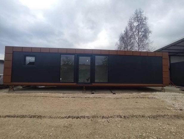 Kontener mieszkalny z płyt warstwowych, czarne ściany, brązowe obramowanie, ustawiony na podporach na żwirowym podłożu, widoczne okno i drzwi przesuwne, pochmurne niebo w tle.