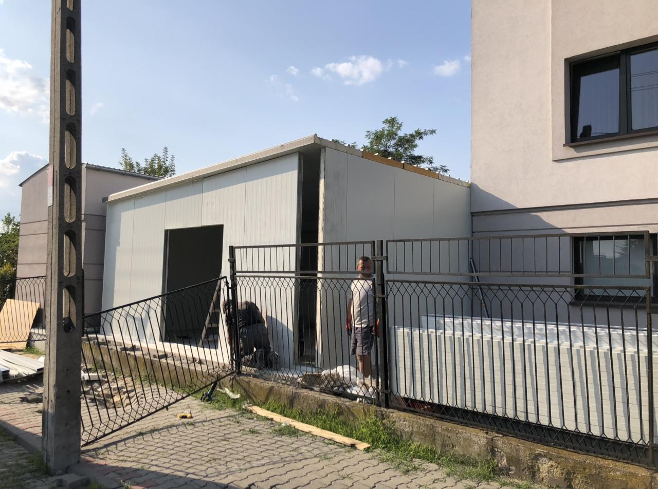 Konstrukcja z płyt warstwowych w trakcie montażu przy budynku mieszkalnym, widoczni dwaj pracownicy, materiały budowlane za ogrodzeniem.