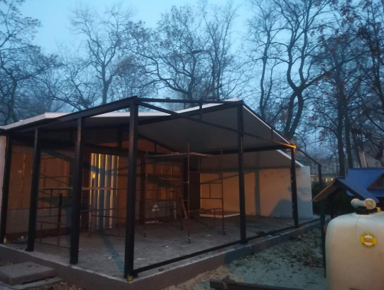 Konstrukcja z płyt warstwowych w trakcie montażu, widoczne rusztowanie i elementy mocujące, tło stanowią drzewa bez liści.