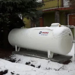 Biały zbiornik na gaz płynny umieszczony na betonowym fundamencie, częściowo przysypany śniegiem, z widocznym logo firmy i numerem telefonu, na tle zimowego ogrodu z domem.