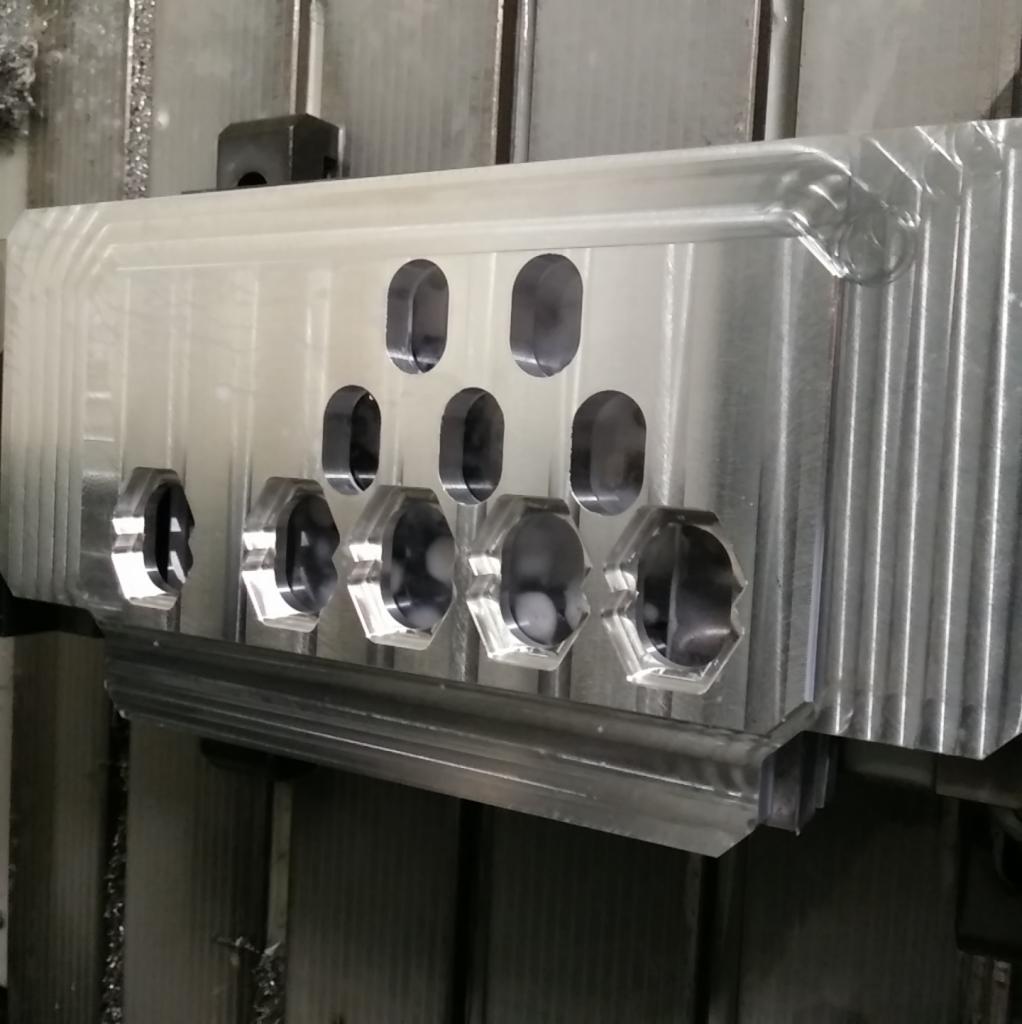 Aluminiowy element po obróbce z widocznymi otworami o różnych kształtach, w tym okrągłych i sześciokątnych, na tle maszyny.