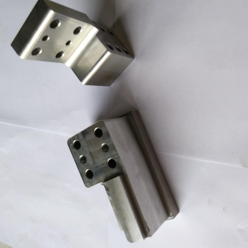 Dwa metalowe, precyzyjnie obrobione elementy w kształcie kątowników z otworami, leżące na białym tle. Jeden element jest ułożony pod kątem, ukazując powierzchnię z sześcioma otworami. Widoczne...