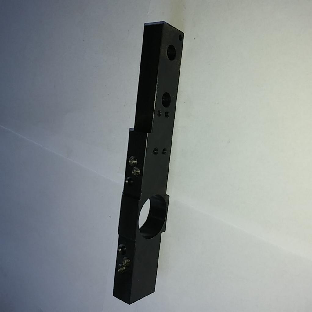 Czarny, metalowy element z otworami o różnych średnicach i gwintami, wykonany z precyzją, leżący na jasnym tle.