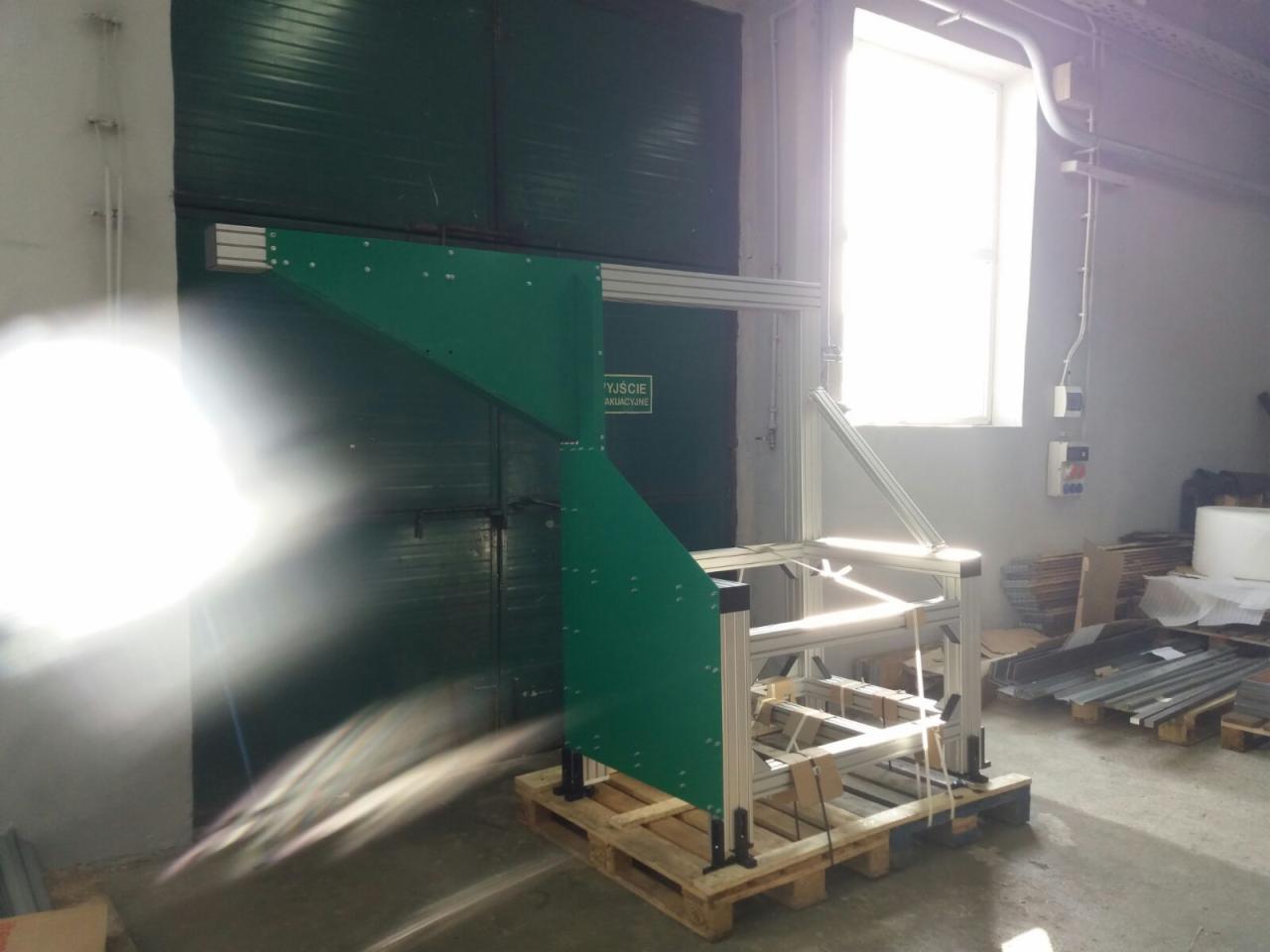 Szkielet konstrukcji z profili aluminiowych i zielonych paneli na palecie, widoczne otwory montażowe, w tle brama przemysłowa i okno z silnym światłem.