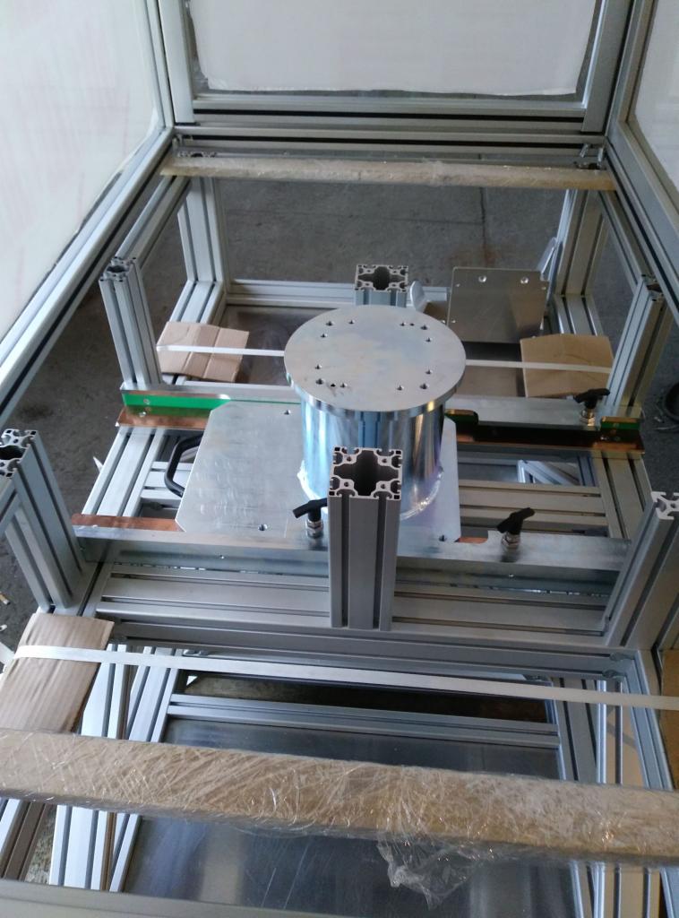 Wewnętrzny widok ramy z profili aluminiowych z zamontowanym cylindrycznym elementem obrabianym, umieszczonym na metalowej płycie z otworami, całość w surowym, warsztatowym otoczeniu.