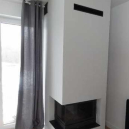 Mar-bud - Nowoczesny, narożny kominek w minimalistycznym stylu, z białym obudowaniem i czarnym wkładem, widoczny z boku, obok okna z szarą zasłoną.