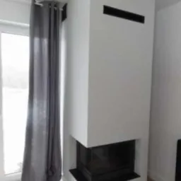 Nowoczesny, narożny kominek w minimalistycznym stylu, z białym obudowaniem i czarnym wkładem, widoczny z boku, obok okna z szarą zasłoną.