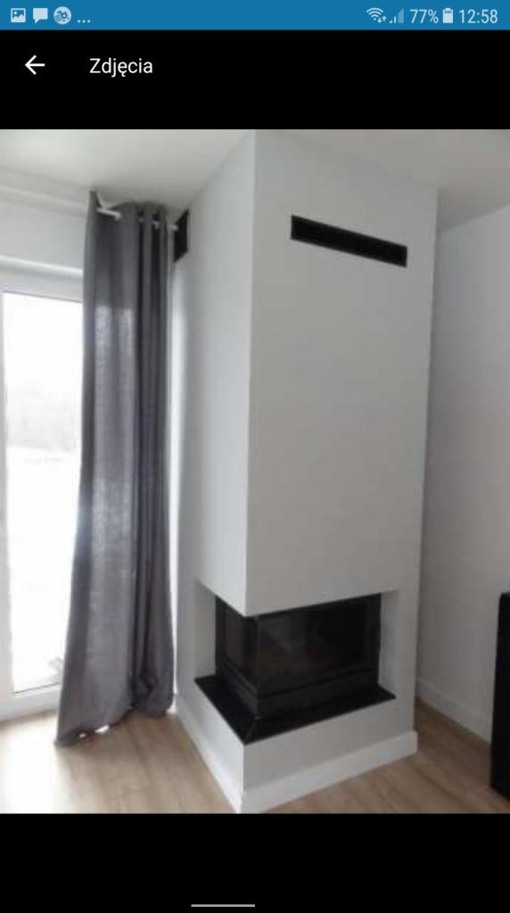 Nowoczesny, narożny kominek w minimalistycznym stylu, z białym obudowaniem i czarnym wkładem, widoczny z boku, obok okna z szarą zasłoną.