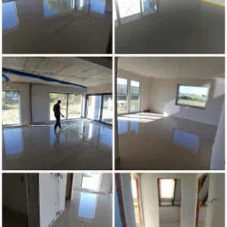 Świeżo wylana, gładka posadzka w surowym wnętrzu, z widocznymi dużymi oknami i odbijającym się w powierzchni niebem. W jednym z kadrów osoba rozprowadza masę poziomującą za pomocą listwy. Żółta...