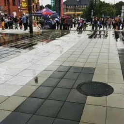 Nowa nawierzchnia placu z szarej kostki brukowej ułożonej w geometryczny wzór, lekko mokra po deszczu, w tle zgromadzenie ludzi i namiot.