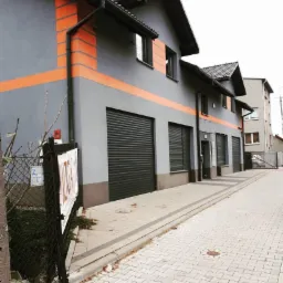 Szary budynek usługowy z pomarańczowymi akcentami i rolowanymi bramami garażowymi, widok z chodnika wyłożonego kostką.