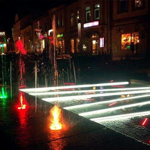 Nocny widok na fontannę z podświetleniem LED w kolorach czerwieni i zieleni, w tle budynki miejskie z oświetlonymi oknami.