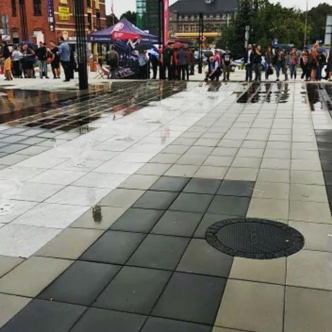 Nowa nawierzchnia placu z szarej kostki brukowej ułożonej w geometryczny wzór, lekko mokra po deszczu, w tle zgromadzenie ludzi i namiot.