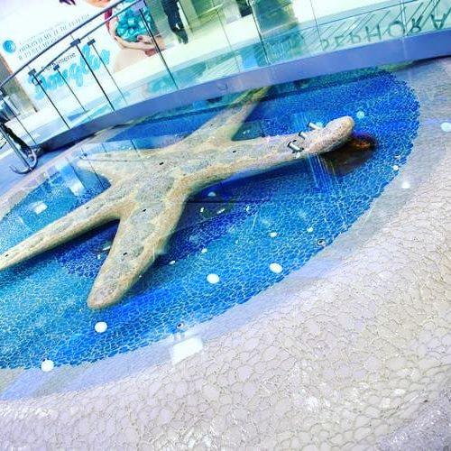 Dekoracyjny element w kształcie rozgwiazdy w płytkim, błękitnym zbiorniku wodnym z białymi kulkami, otoczonym jasną mozaiką w centrum handlowym.