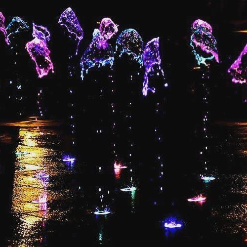Podświetlana fontanna z wieloma strumieniami wody w kolorach fioletu, różu i błękitu, widoczna nocą, tworząca refleksy na mokrej powierzchni.