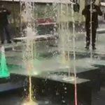 Dysze fontanny podświetlone kolorowymi światłami, tryskające wodą na czarnej, kwadratowej powierzchni. W tle widoczne sylwetki ludzi.