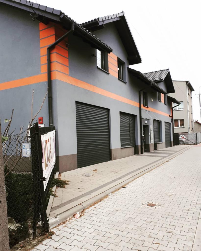 Szary budynek usługowy z pomarańczowymi akcentami i rolowanymi bramami garażowymi, widok z chodnika wyłożonego kostką.