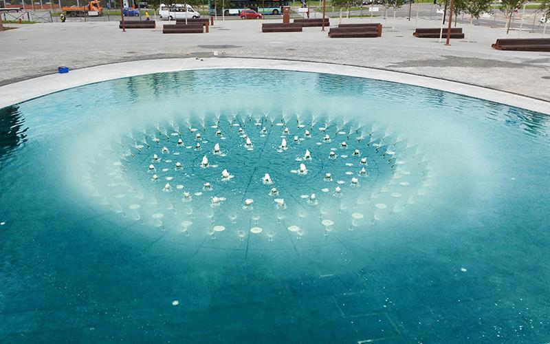 Okrągła fontanna z wieloma dyszami tryskającymi wodą, otoczona betonowym placem z drewnianymi ławkami w tle, w pobliżu budynku.