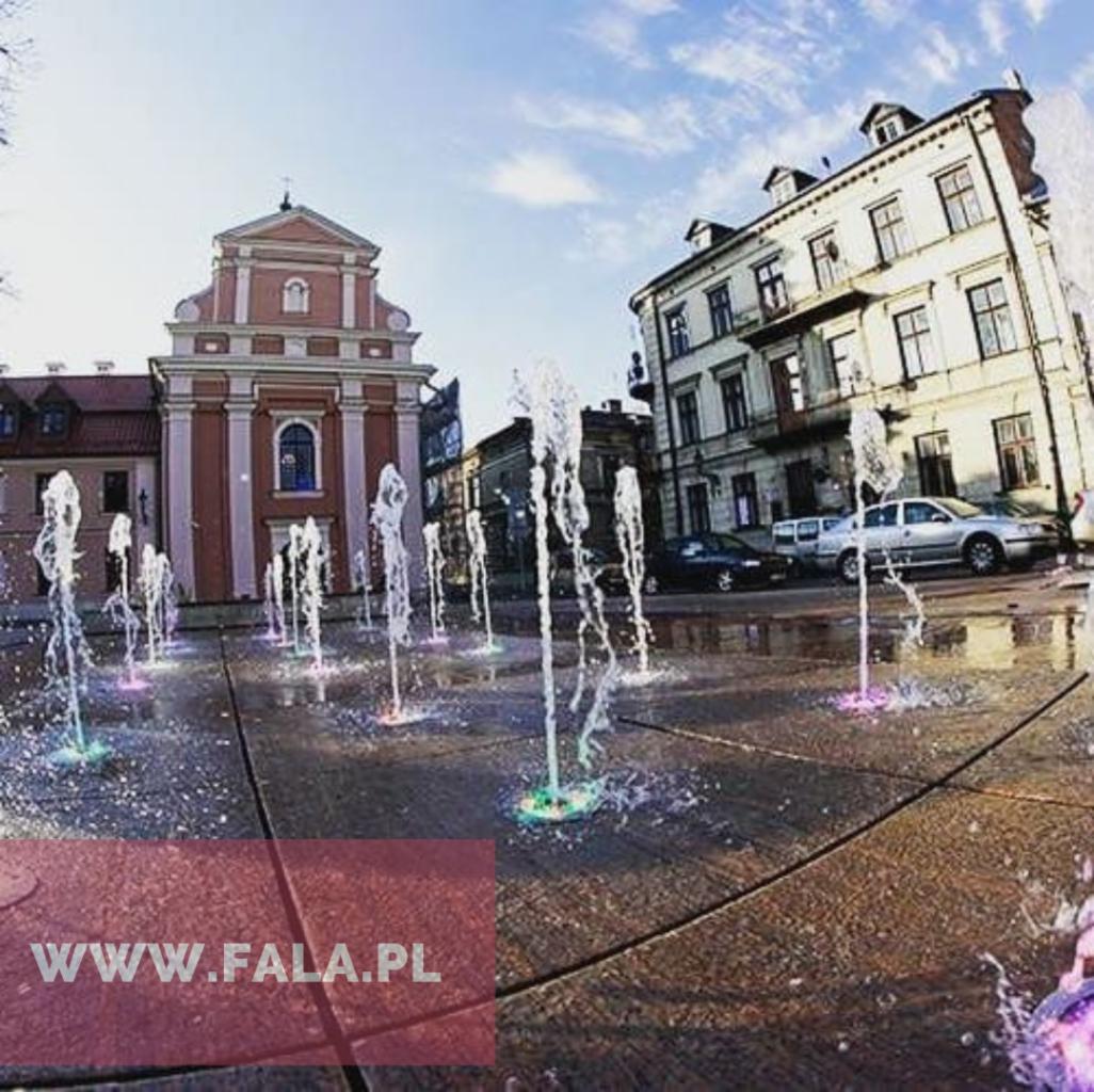 Plac miejski z fontanną podświetlaną kolorowymi diodami, w tle zabytkowe budynki i kościół w słoneczny dzień.