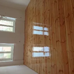 Ściana wykończona jasnymi panelami drewnianymi z widocznymi refleksami okien, świeżo po montażu, w narożniku pomieszczenia z dwoma oknami.