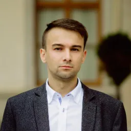 Portret młodego mężczyzny w szarej marynarce i białej koszuli z wzorzystym kołnierzykiem, na rozmytym tle budynku i zieleni.