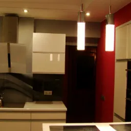 Nowoczesna kuchnia w odcieniach szarości i bieli z akcentami czerwieni, wyposażona w białe szafki, szklany panel nad blatem z motywem fali i wiszące lampy o cylindrycznym kształcie.