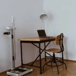 W gabinecie: waga medyczna z analizatorem składu ciała, biurko z czarnymi nogami i drewnianym blatem, laptop oraz lampa podłogowa na tle białej ściany.
