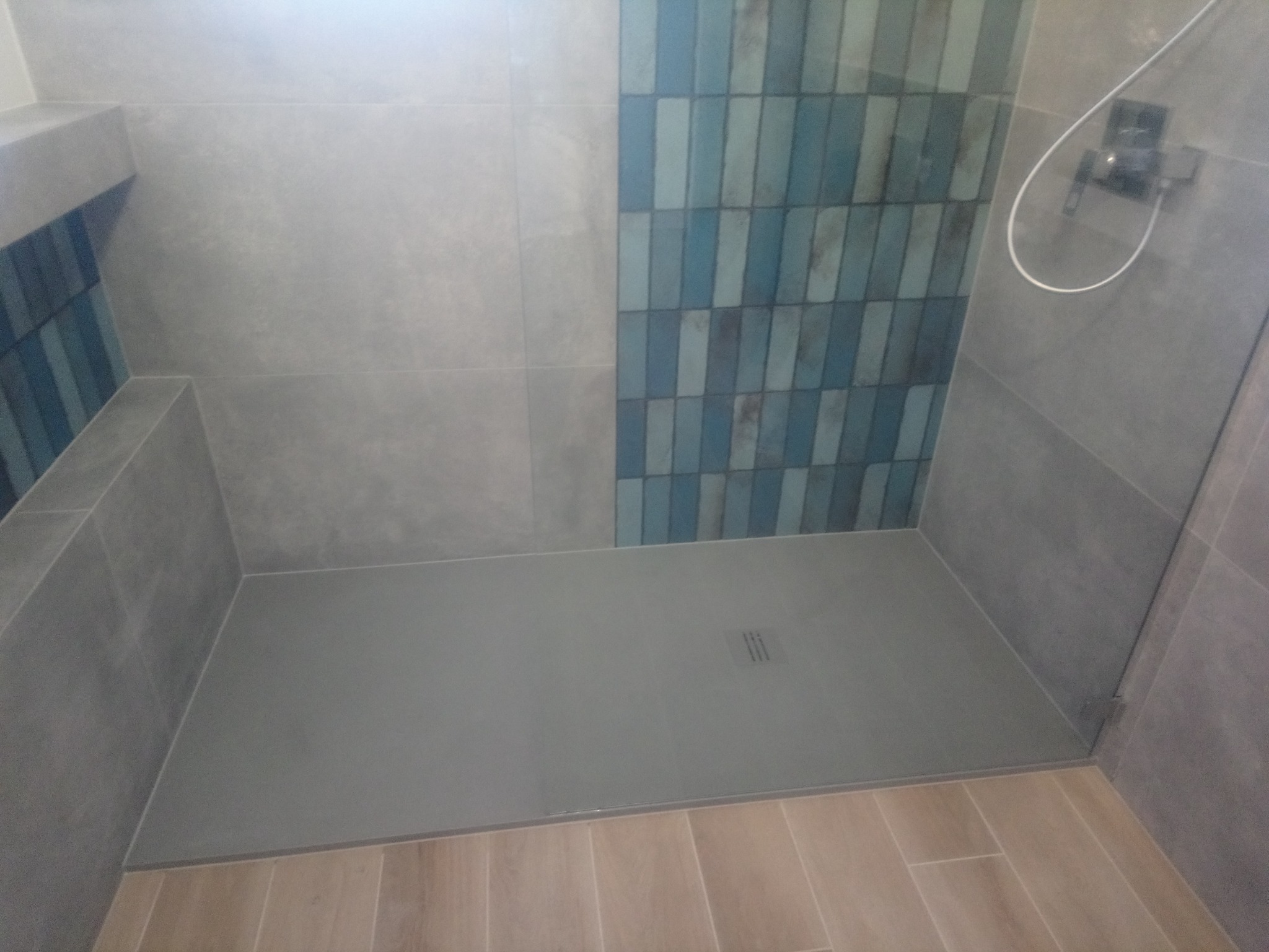 Wykończona kabina prysznicowa z szarymi płytkami i niebieską mozaiką na ścianie, szary brodzik z odpływem liniowym, jasne panele podłogowe.