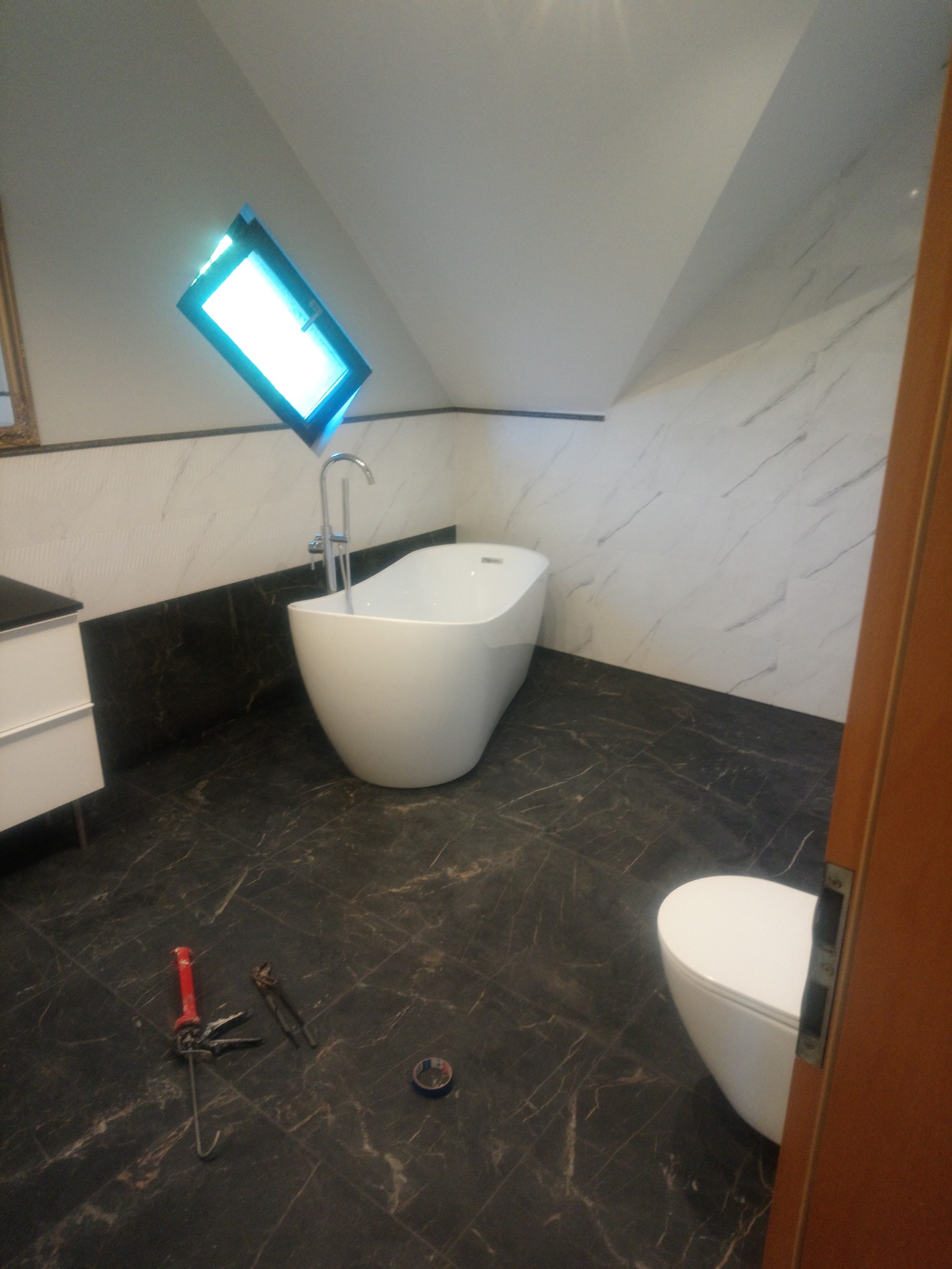 Nowoczesna łazienka na poddaszu z wolnostojącą wanną, marmurowymi płytkami ściennymi i ciemną podłogą; na podłodze leżą narzędzia glazurnicze.