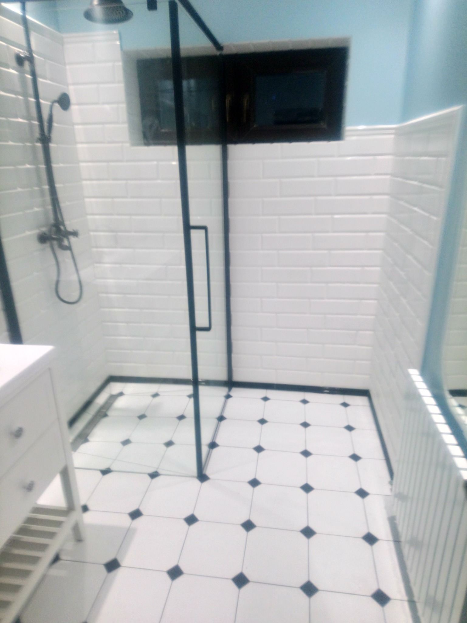 Nowoczesna kabina prysznicowa z czarnymi elementami, białe płytki typu metro na ścianach i podłodze z białych płytek z czarnymi akcentami, widoczna umywalka z szafką w stylu klasycznym.