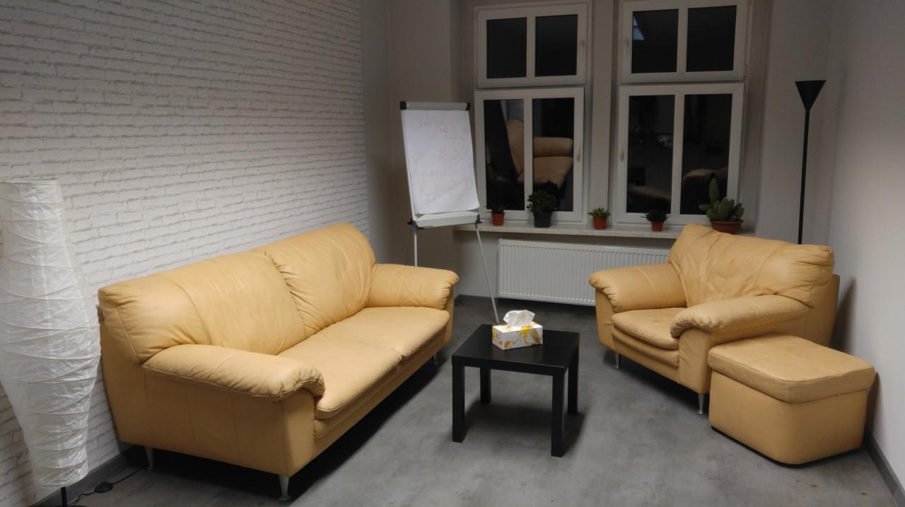 Jasny gabinet z beżową sofą, fotelem z podnóżkiem, czarnym stolikiem z chusteczkami, flipchartem i lampą podłogową w rogu, okno z roślinami.