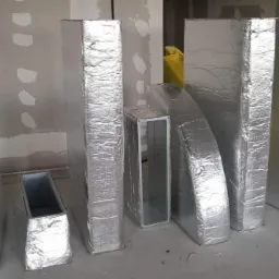 Izolowane kanały wentylacyjne o przekroju prostokątnym i okrągłym, ustawione na podłodze w surowym wnętrzu, przed montażem.