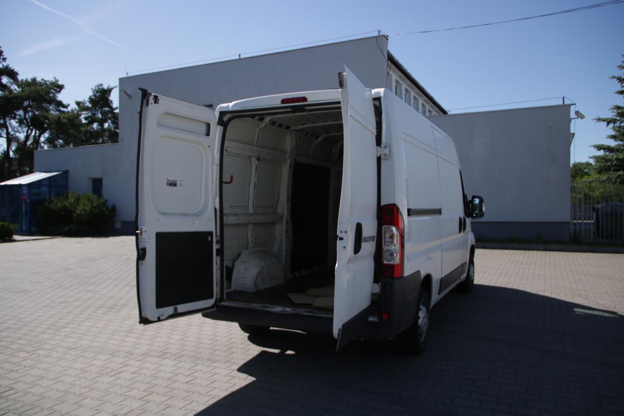 Biały bus z otwartymi tylnymi drzwiami, widok z tyłu na puste wnętrze, zaparkowany na brukowanym placu przed budynkiem.