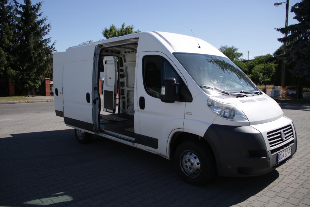 Biały furgon z otwartymi drzwiami bocznymi i tylnymi, ukazujący puste wnętrze ładunkowe, zaparkowany na brukowanej powierzchni w słoneczny dzień.