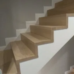 Jasne drewniane schody z białymi listwami przypodłogowymi i szarą ścianą w tle.