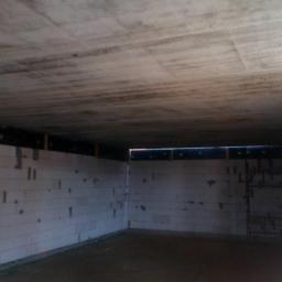 Krystian Szukalski - Surowe wnętrze budynku w trakcie budowy, ściany z białych bloczków, betonowy sufit z widocznymi śladami szalunków, metalowe zbrojenie w ścianie po prawej stronie.