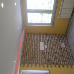 Krystian Szukalski - Wnętrze pokoju z dekoracyjną ścianą z cegieł, żółtą ścianą, grzejnikiem i podłogą imitującą drewno, oświetlenie punktowe w suficie.