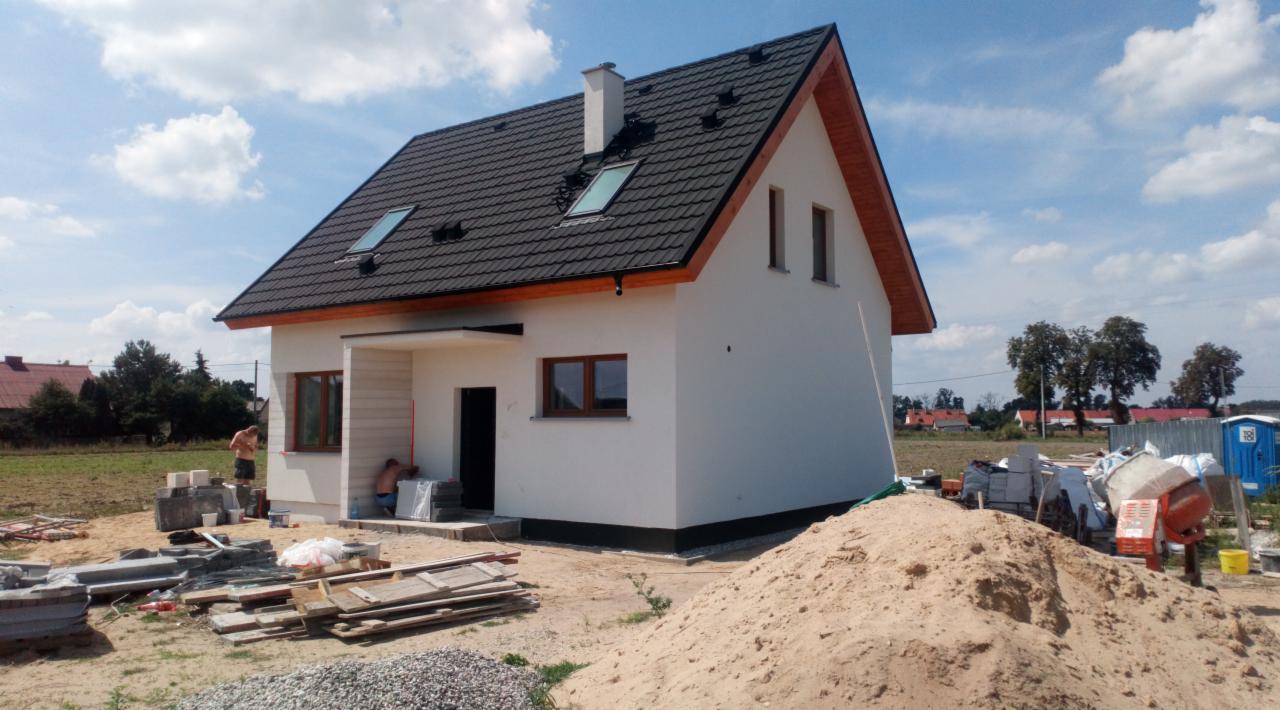 Nowo wybudowany dom jednorodzinny z białą elewacją, czarną dachówką i drewnianym wykończeniem dachu, widoczne prace wykończeniowe wokół budynku, w tym materiały budowlane, piasek i betoniarka.