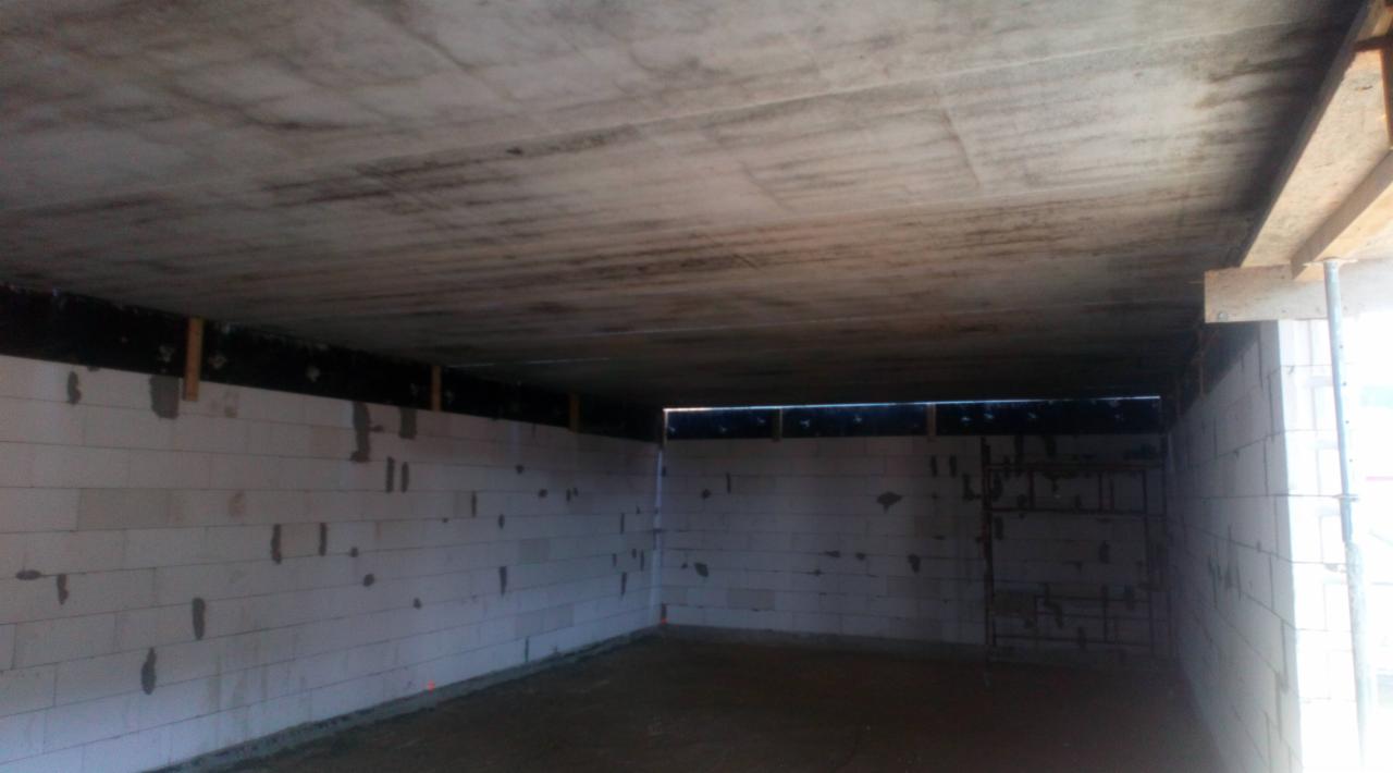 Surowe wnętrze budynku w trakcie budowy, ściany z białych bloczków, betonowy sufit z widocznymi śladami szalunków, metalowe zbrojenie w ścianie po prawej stronie.