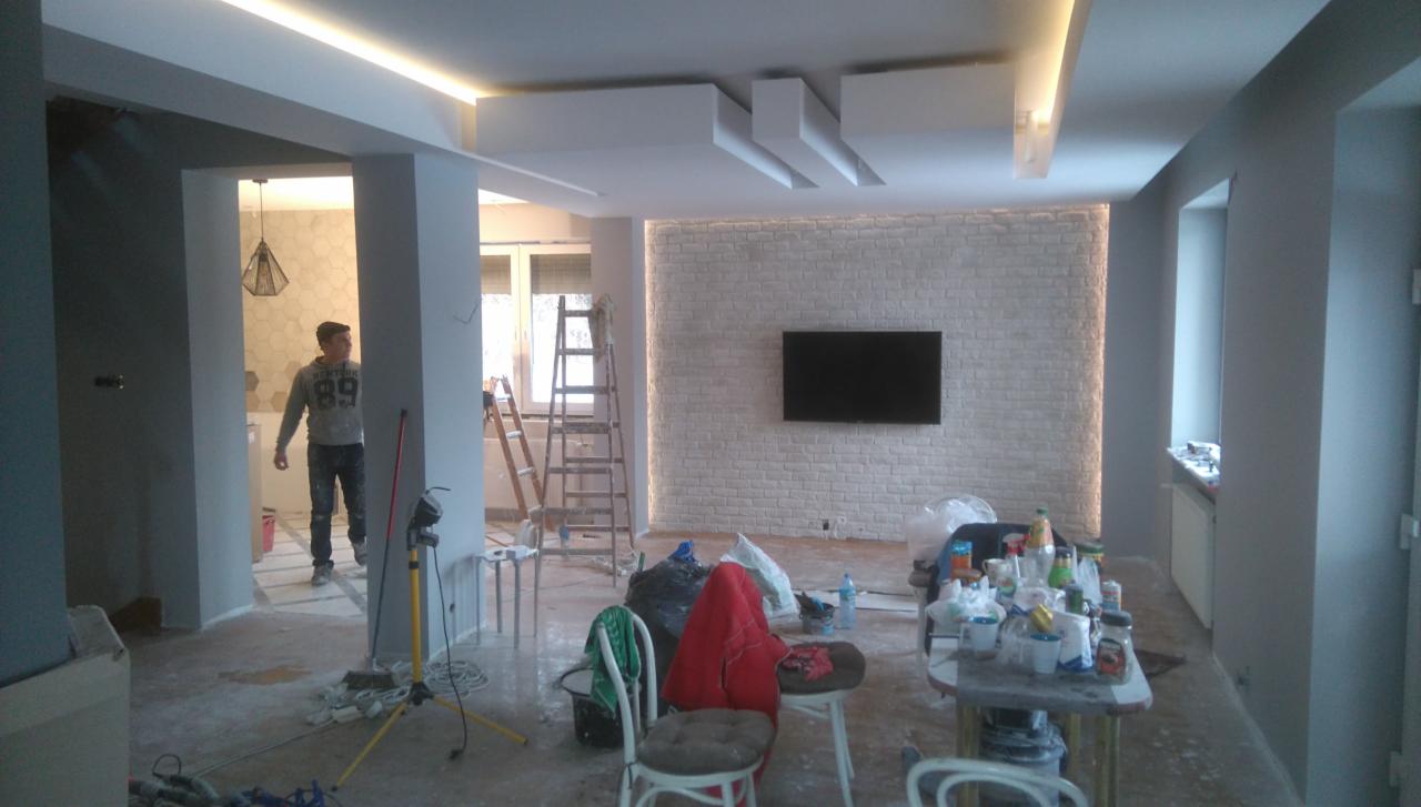 Wnętrze w trakcie wykańczania w Przygodzicach: ściana z białej cegły z podświetleniem LED, widoczny pracownik, rozstawione drabiny i narzędzia, podłoga zabezpieczona folią.