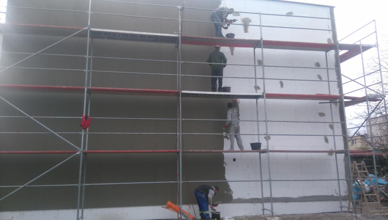 Prace na elewacji budynku z użyciem rusztowania, widoczni pracownicy podczas nakładania tynku na ocieploną ścianę, fragment otynkowanej powierzchni oraz niezabezpieczona drabina oparta o rusztowanie.