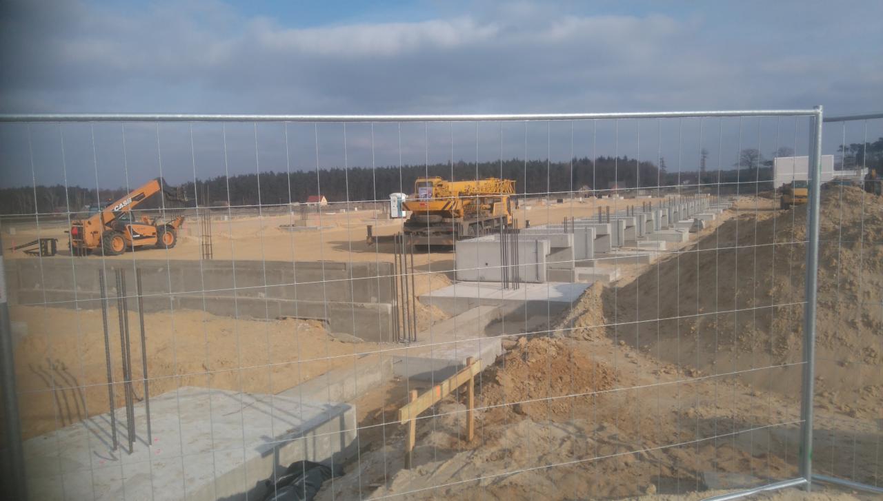 Fundamenty budynku w trakcie budowy, widoczne zza ogrodzenia placu budowy, z dźwigiem i podnośnikiem teleskopowym w tle.