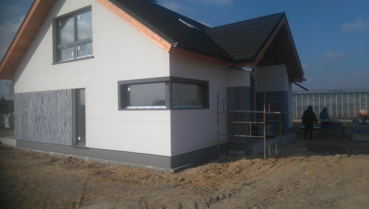 Nowoczesny dom jednorodzinny z elewacją w kolorze białym i szarym, z widocznymi fragmentami rusztowania i pracującymi osobami w tle.