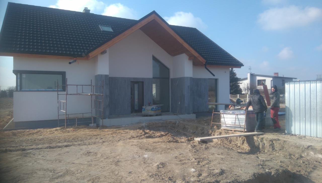 Nowoczesny dom jednorodzinny w trakcie wykończenia elewacji, z widocznymi szarymi panelami dekoracyjnymi, rusztowaniem i dwoma pracownikami na zewnątrz.