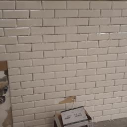 Mirex Bud - Ściana w trakcie układania białych, prostokątnych płytek w stylu metro, z widocznymi krzyżykami dystansowymi i częściowo odsłoniętą instalacją hydrauliczną po lewej stronie.