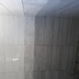 Ściana wyłożona jasnymi i ciemnymi płytkami imitującymi drewno, z dekoracyjnym pasem płytek w kwiatowy wzór.