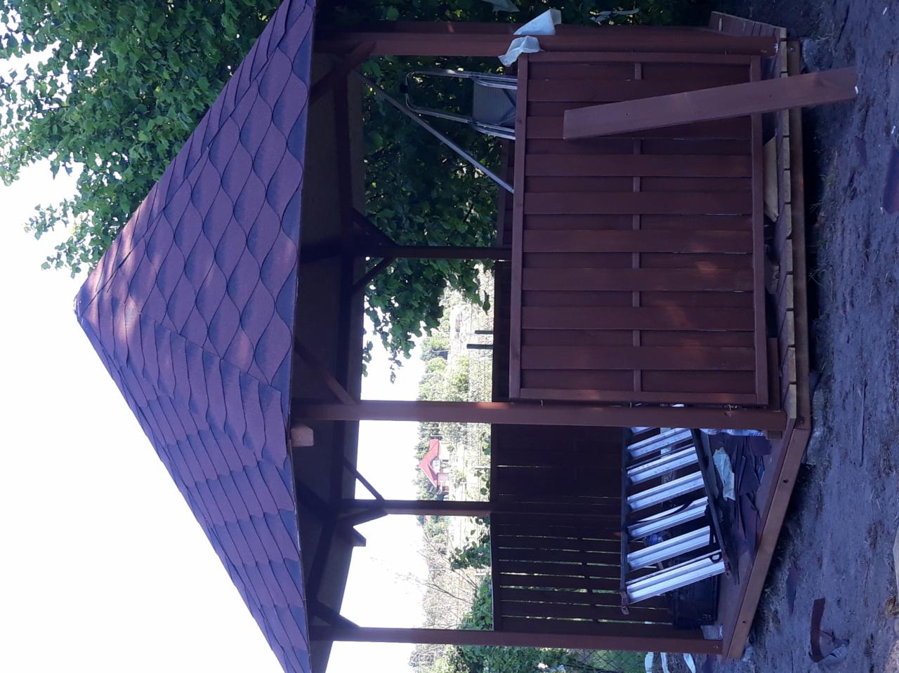 Drewniana altana ogrodowa z bordowym dachem bitumicznym w trakcie budowy, widoczne deski, drabina i narzędzia.