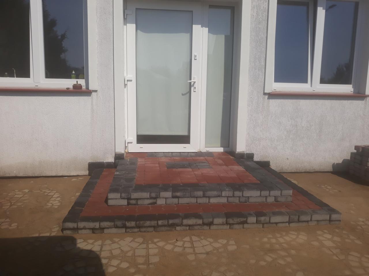 Schody wejściowe do domu wyłożone kostką brukową w kolorach czerwonym i szarym, z widoczną fakturą kostki i fugami, otoczone piaskowym podłożem.
