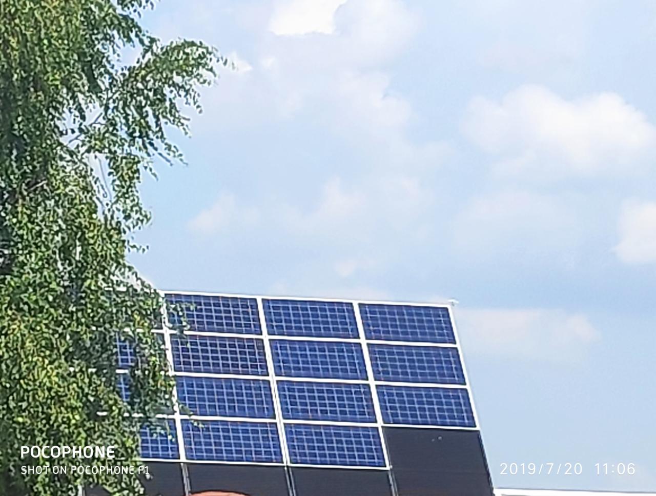 Panele fotowoltaiczne na dachu budynku, częściowo zasłonięte przez drzewo. Błękitne niebo z białymi chmurami w tle. Zdjęcie z datą w prawym dolnym rogu.